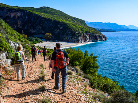 Hikers walking a rocky trail above the Albanian Ionian Coast, with the turquoise sea and hidden coves below