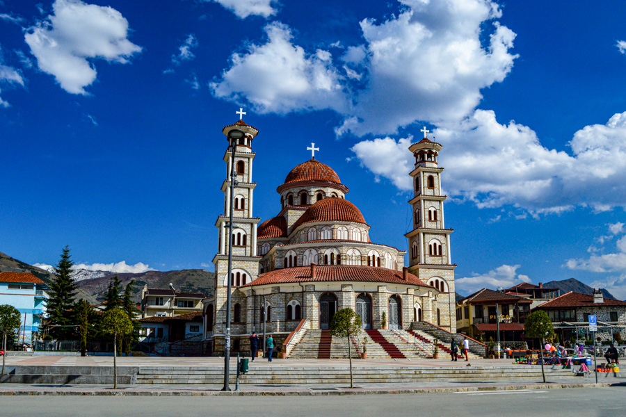 Resurrection Cathedral in Korçë with red-domed roof and twin bell towers, seen from the main square under a bright blue sky.